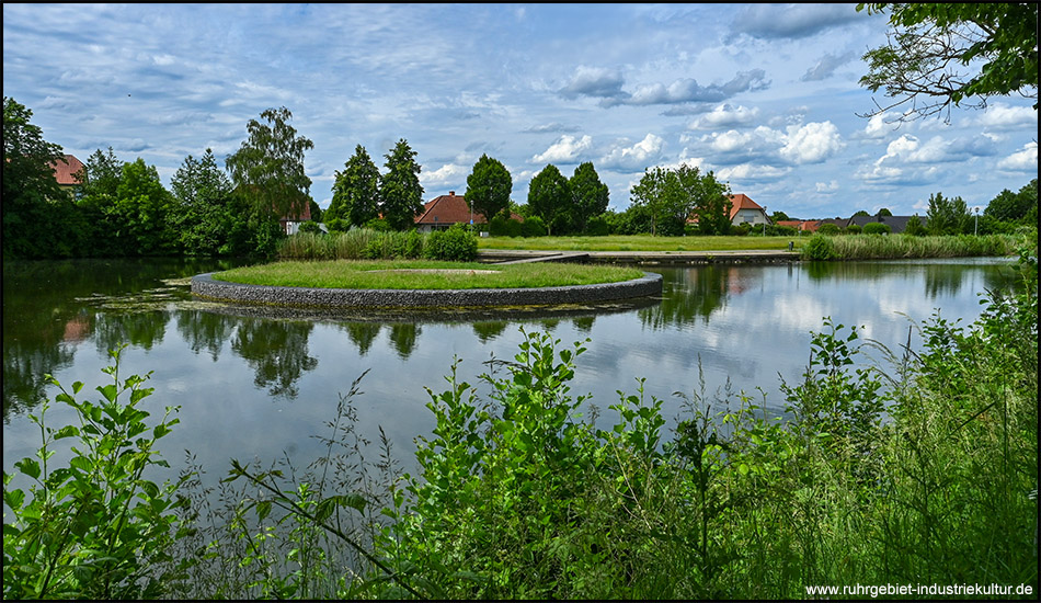 Ein Teich mit einer runden künstlichen Insel. Im Hintergrund sind Häuser einer Ortschaft zu sehen