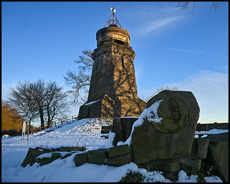 Von der Sonne beleuchteter Bismarckturm mit Schnee auf der Fläche. Im Vordergrund ist eine Plakette mit dem Bismarck-Portrait, das teilweise verschneit ist.