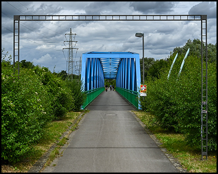 Radweg führt auf eine blaue Stahlfachwerkbrücke zu