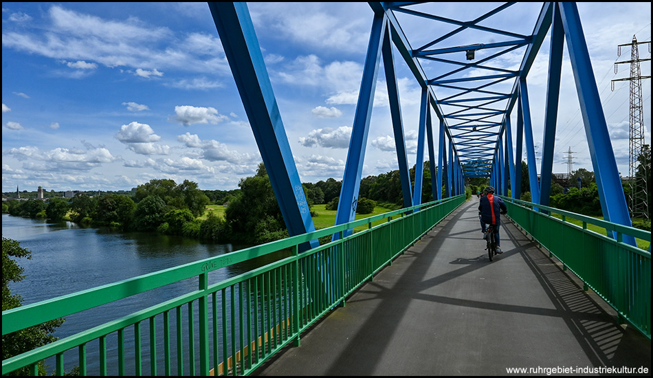 auf einer Brücke auf einem Fluss. Sie wird von einem blauen Stahlfachwerk getragen und von grünen Geländern begrenzt.