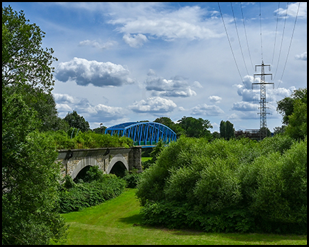 Blick über eine Wiese zu einer Brücke mit blauem Fachwerkträger