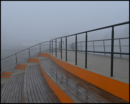 Stufen als Sitzgelegenheiten aus Holz und orangefarbener Rückenlehne. Teil einer Brückenanlage im Nebel