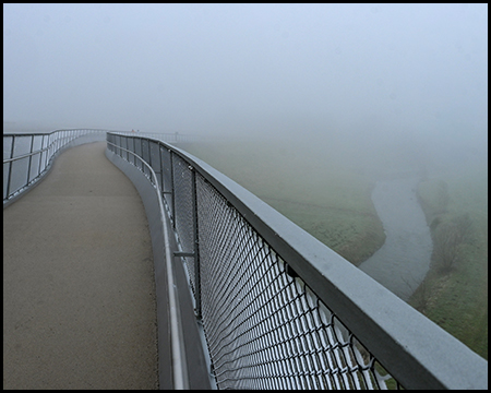 Ein Gehweg auf einer Brücke über einem Fluss. Die Szene ist im dichten Nebel.