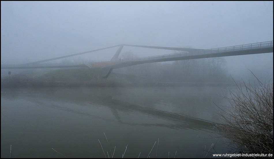 Eine geschwungene Brücke über einen Kanal im Nebel. Zu sehen ist schwach ein geneigter Pylon, der Teile der Brücke trägt.