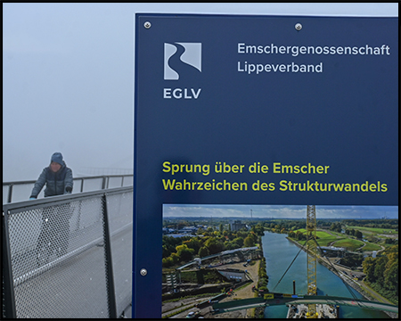 Ein Radfahrer auf einem Brücken-Weg. Daneben dominant eine Erläuterungstafel der Emschergenossenschaft zur Brücke "Sprung über die Emscher", Wahrzeichen des Strukturwandels