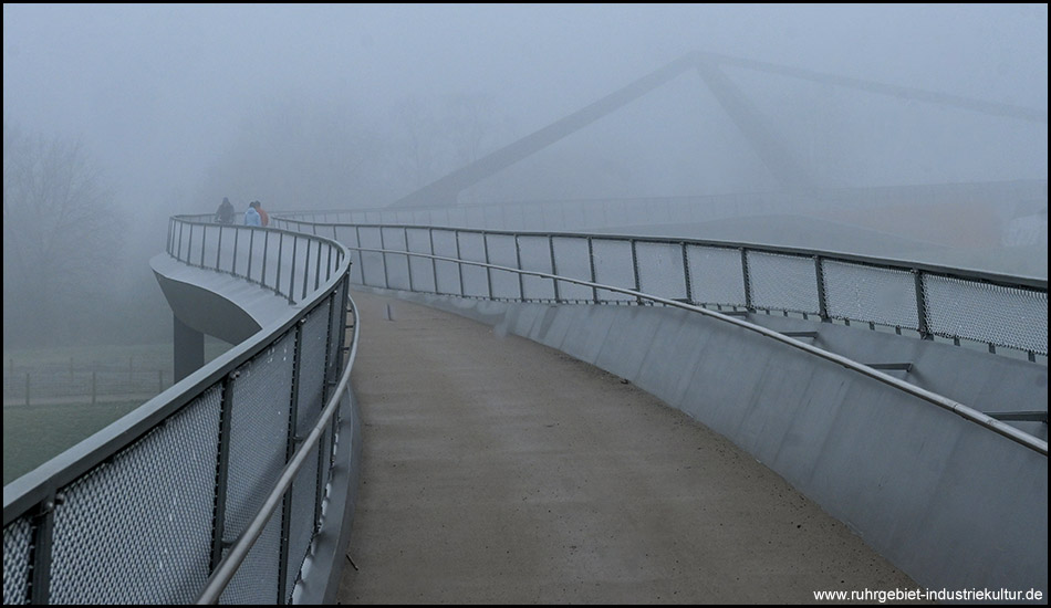 Kurvenförmiger Fußweg auf einer Brückem die in einen dichten Nebel führt. Bauteile der Brücke wie ein Brückenpylon sind im Nebel.