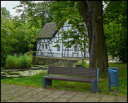 Eine Bank unter einem Baum vor einem Teich mit Fachwerkhaus