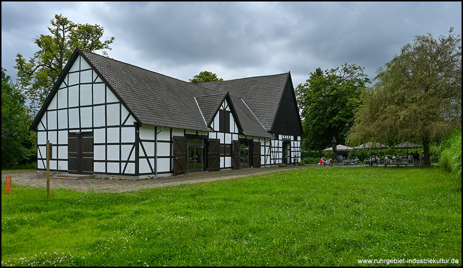 Fachwerkgebäude mit einer Wiese davor und einem alten Baum. Angedeutet sichtbar sind Tische und Stühle einer Gastronomie.