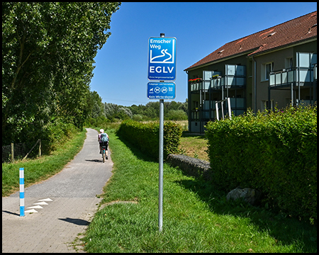 Schild vom Emscherweg am Rande eines Radwegs neben einem Wohnhaus