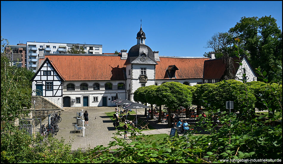 Ein altes Wasserschloss mit einem markanten Turm mit Turmhaube. Davor Bäume in einer Parkanlage