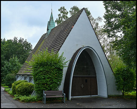 Eine kleine Friedhofskapelle. Der Eingang ist ein Parabelbogen und die Kapelle besteht nur aus einem Zeltdach.