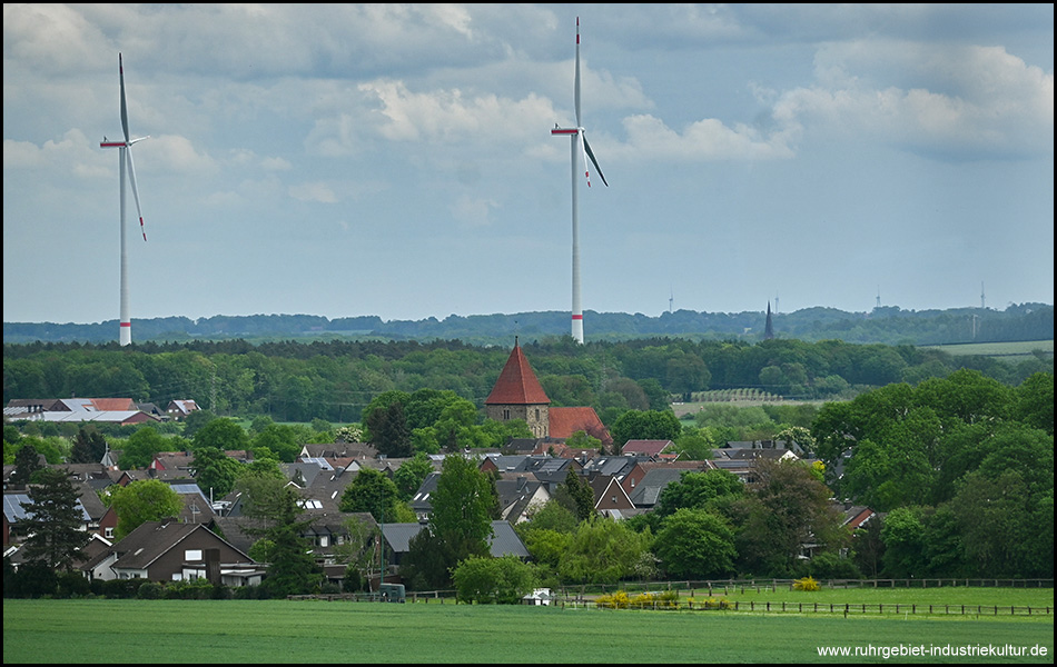 Ausblick über Felder auf eine Siedlung mit markanter alter Kirche und zwei Windrädern am Horizont