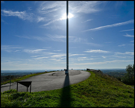 Gegenlichtbild mit dem Gipfel eines Berges, auf dem eine schmale, 30 Meter hohe Skulptur  genau im Gegenlicht der Sonne steht