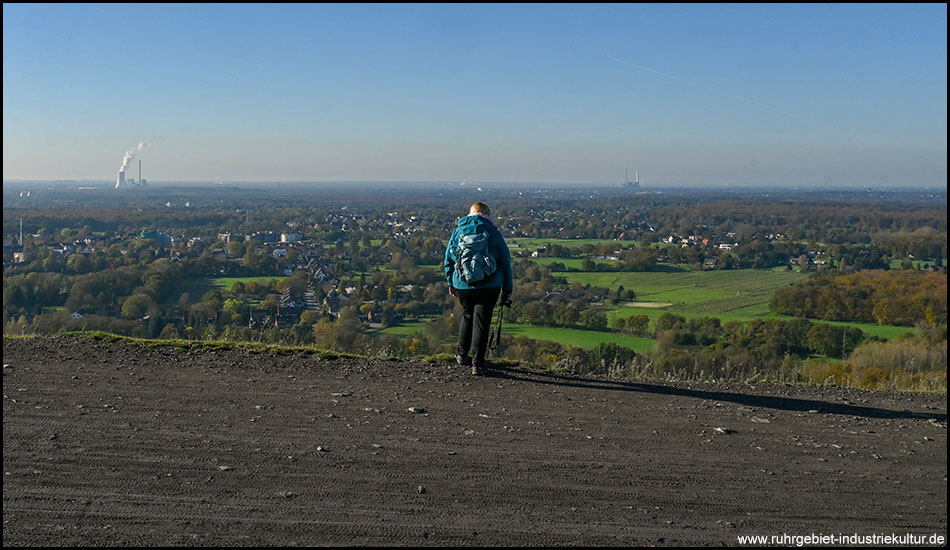 Eine Frau steht vor dem Abgrund an einem Berg und blickt in die Ebene davor. Hier sind Bäume, Wälder, Hecken und Siedlungen zu sehen. Im Hintergrund ein Kraftwerk mit Kühlturm.