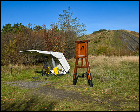 Schildausbau aus dem Bergbau als ausgestelltes Objekt, daneben ein Schild in Förderturm-Optik mit Bibelzitat als Teil des Kreuzwegs. Im Hitnergrund Hügelkuppe als Gipfel der Halde Haniel