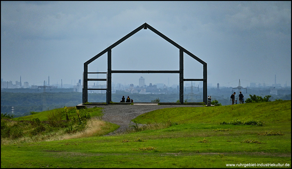 Giebelseite des Gerüstes eines Hauses auf einem Berggipfel. Im Hintergrund ist eine Kulisse von Industrie und Gebäuden am Horizont zu sehen