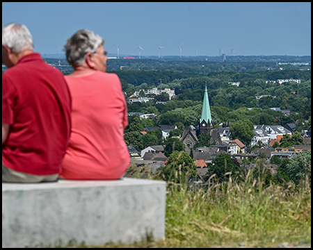Zwei ältere Personen schauen auf einem Betonquader sitzend auf einem Hügel hinab auf einen Ort mit einer Kirche