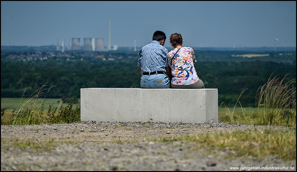 Ein Paar aus Mann und Frau sitzt auf einem Betonquader auf dem Gipfel einer Halde. Im Hintergrund grüne Ebene und ein einzelnes Kraftwerk