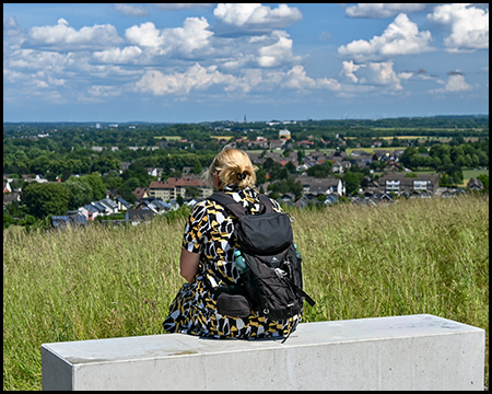 Eine Frau sitzt auf einem Sitzquader aus Beton auf dem Gipfel der Halde Schleswig und schaut in die Ferne.