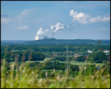 Blick durch unscharfe Gräser auf einem Gipfel einer Halde auf die Umgebung mit einem Kraftwerk und einer Bergehalde