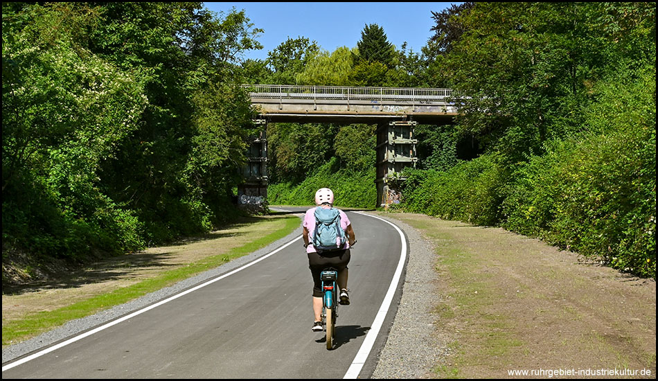 Eine Radfahrerin auf einem asphaltierten breiten Radweg kurz vor einer Brücken-Überführung