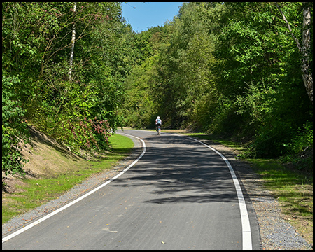 Ein asphaltierter Radweg in einer langen Linkskurve zwischen bewachsenen Böschungen