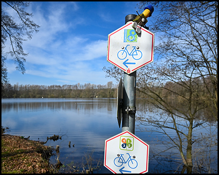 Ein Radweg-Schild vor einem See