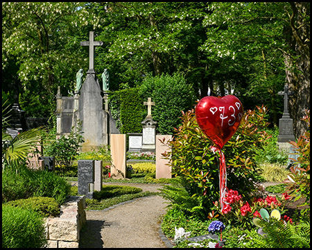 Ein Friedhof mit Grabsteinen. an einem Grab steigt ein Heliumballon in Form eines Herzens auf