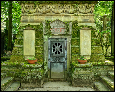 Eine monumentale Gebäude-Gruft mit bemooster Wand und einer über zwei Treppen tiefer erreichbare Tür mit Grünspan