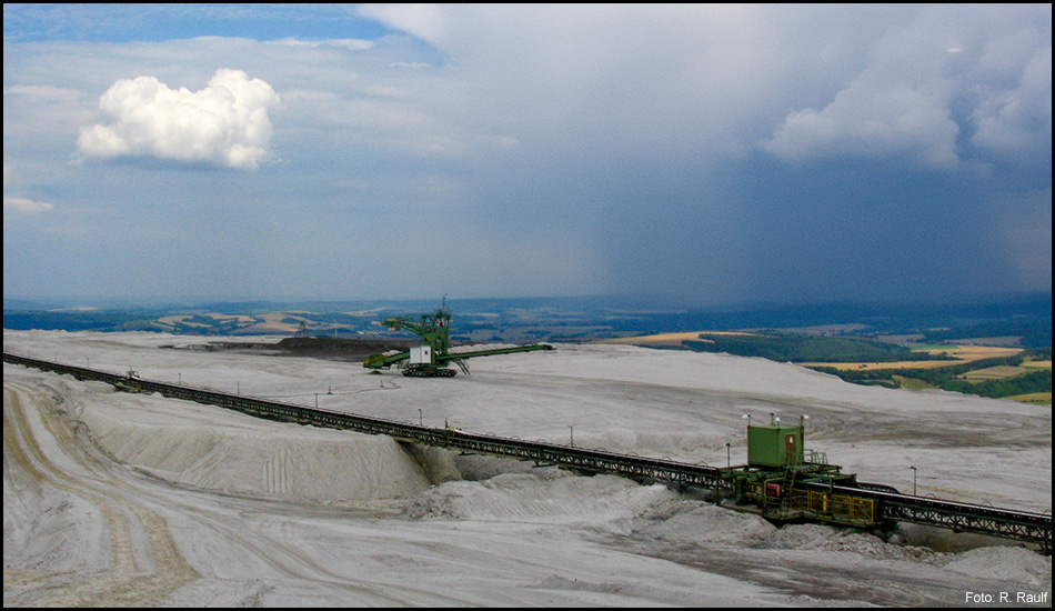 Szene vom Gipfel einer Halde mit Baggern und Transportgeräten sowie Aussicht in die Umgebung mit einem Regenschauer am Horizont