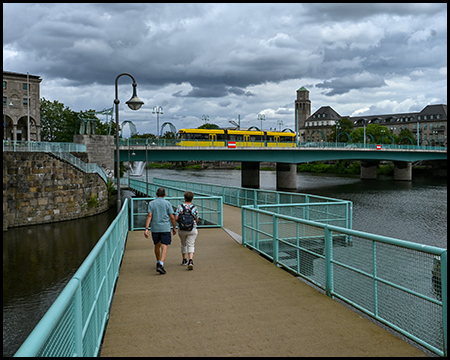 Ein Steg führt auf eine Brücke zu, die quer über einen Fluss verläuft. Darüber fährt eine Straßenbahn