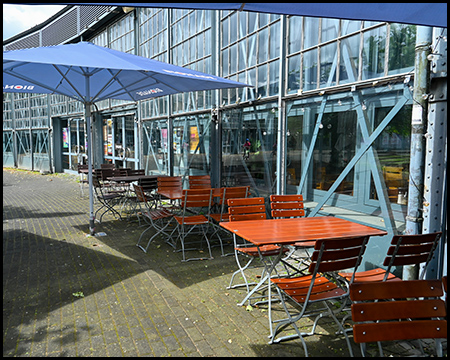 Stühle und Tische einer Außengastronomie vor einem halbrunden Gebäude mit viel Stahl und Glas
