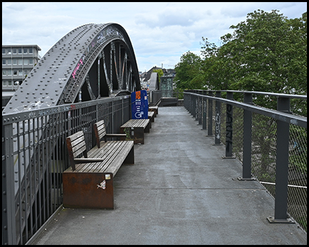 Fußweg mit Bänken vor einem markanten Stahlfachwerkbogen auf einer Brücke