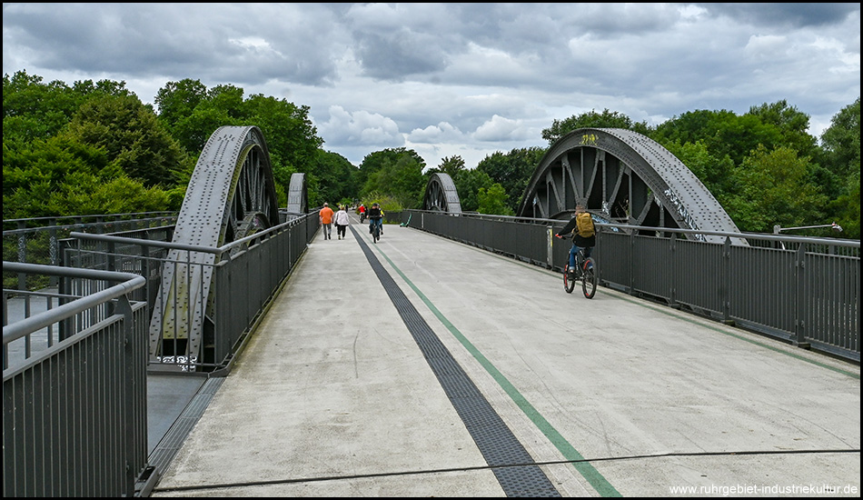 Fahrbahn mit Fuß- und Radweg auf einer Brücke mit stählernen Brückenbögen rechts und links