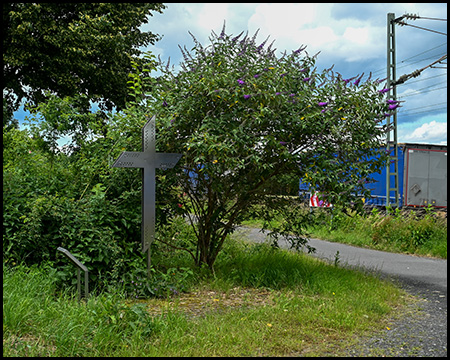 Stählernes Kreuz am Rande eines Weges. Dahinter fährt gerade ein Zug vorbei