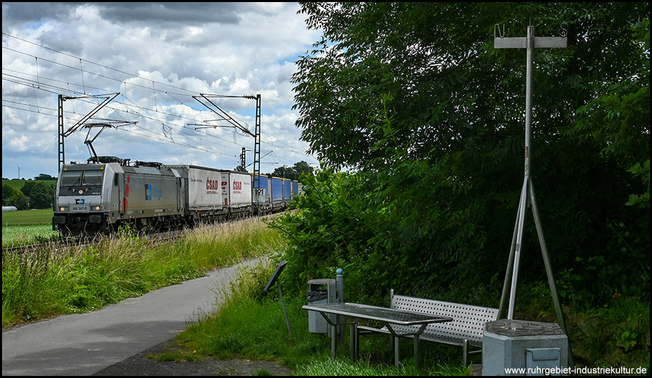 Ein Zug fährt auf einer Bahnstrecke neben einem asphaltierten Wanderweg. Am Rand befindet sich auf der anderen Seite eine Sitzbank mit einer Skulptur, die den Mittelpunkt der Stadt Unna darstellt