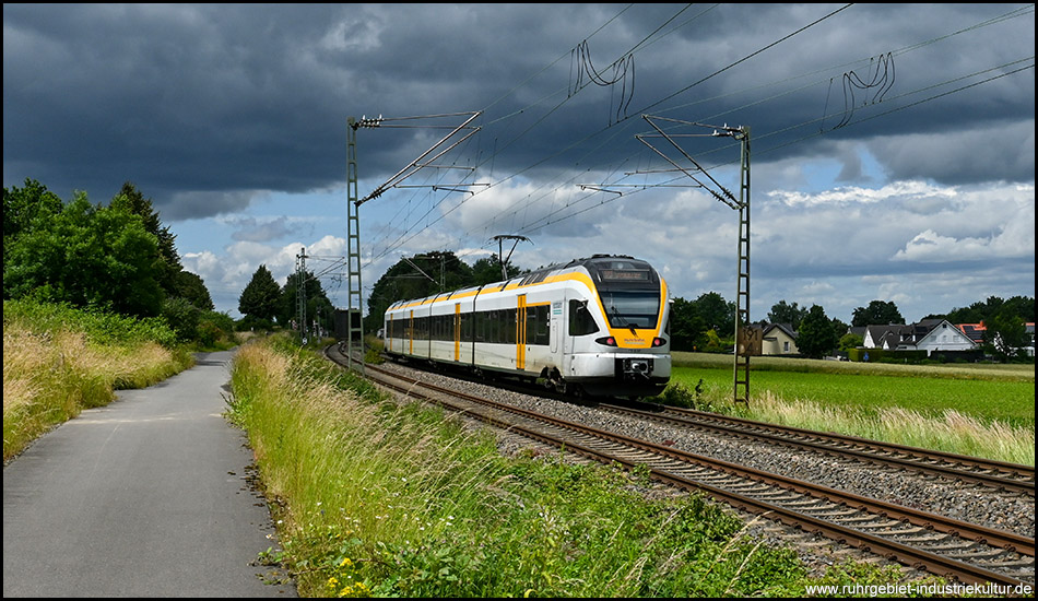 Ein gelb-weißer Triebwagen der eurobahn fährt auf einer Eisenbahnstrecke vor düsterem Himmel parallel zu einem Wirtschaftsweg