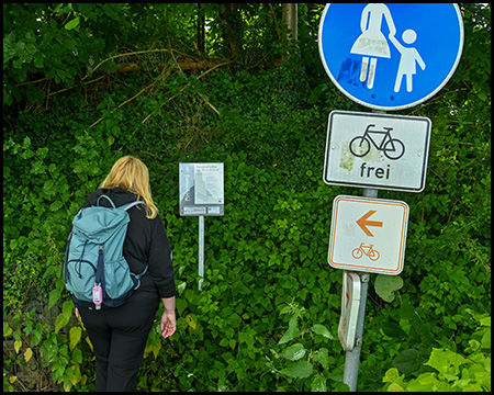 Eine Person mit Rucksack vor einer Infotafel zum Poesieweg an einer Böschung. Daneben ein Schild, das einen Fußgängerweg markiert sowie Schilder für Radverkehr