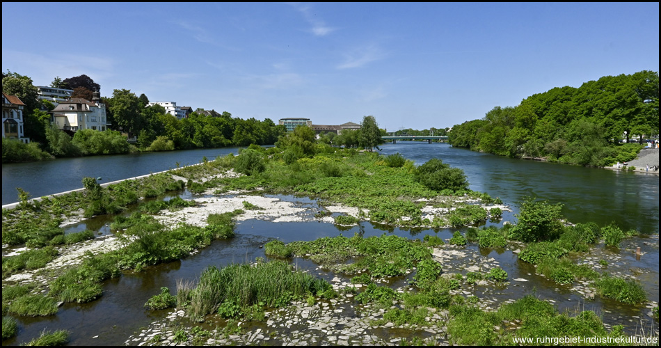 Fluss mit zwei Fließen und einem längs verlaufenden Stauwehr. In der Mitte eine Art Insel mit Geröll und Pflanzen