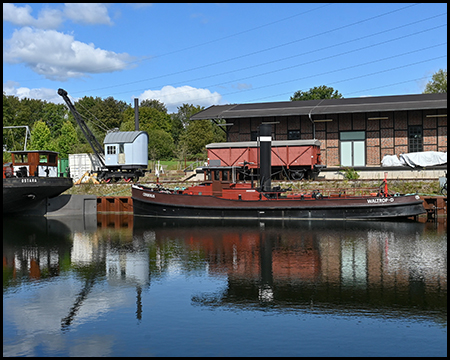 Schiffe am Ufer eines Kais spiegeln sich mit einem Kran und einem Eisenbahnwaggon im Wasser eines Kanals