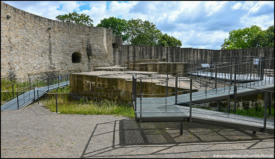 Mauerreste von Türmen und Gebäuden innerhalb einer Ringmauer einer Burg. Ein Steg führt auf Mauerreste des Bergfrieds hinauf