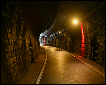 Ein Tunnel mit einem Radweg, der beleuchtet ist. Der Tunnel ist gekrümmt. Licht am anderen Ende ist in der Kurve zu erahnen