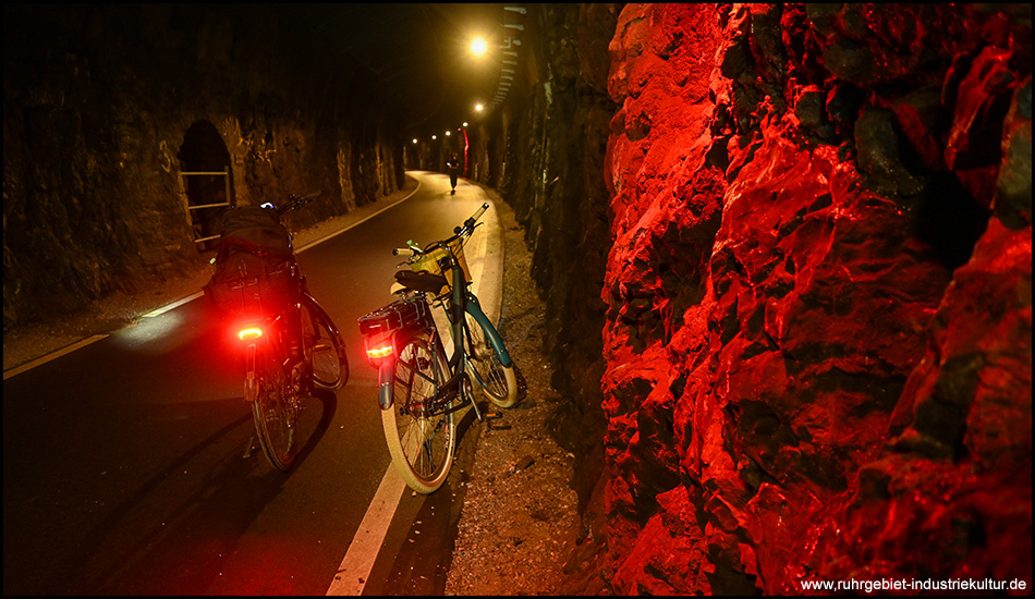 Zwei Fahrräder parken am Rand eines Weges in einem Tunnel. An dieser Stelle ist dieser an der Wand rot beleuchtet. Hier sieht man teils nackten Fels. Die Fahrräder fallen mit ihrer Beleuchtung im Dunkeln auf.