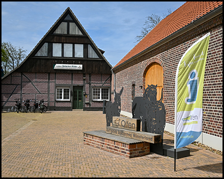 Zwei Gebäude. Davor eine Skulptur zum Thema Münsterland. Eine Fahne mit einem Informations-i