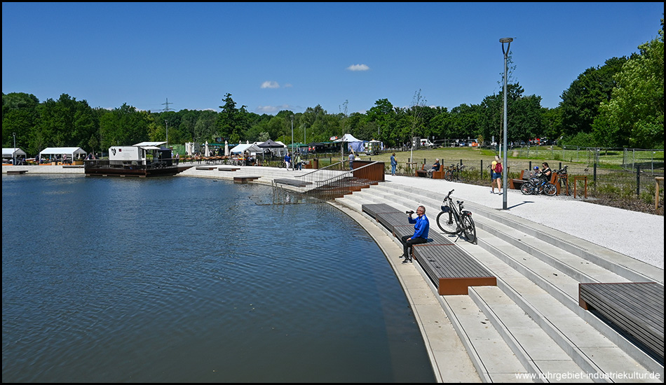 Ein Radfahrer sitzt auf einer Bank am Ufer eines Sees im Bereich einer Promenade