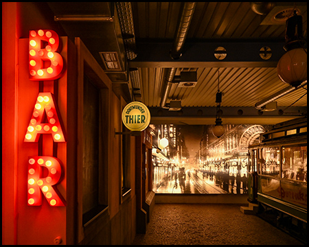 Leuchtende Glühlampen bilden das Wort BAR. Dahinter ein Bild einer nächtlichen Straßenszene